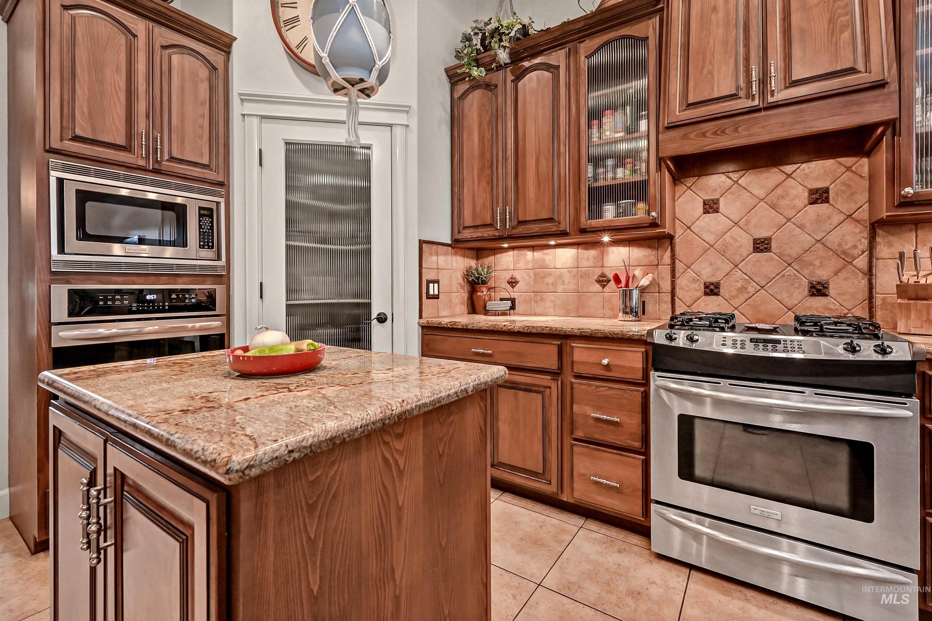 Kitchen with stainless steel appliances, backsplash, brown cabinets, light stone countertops, and light tile patterned floors