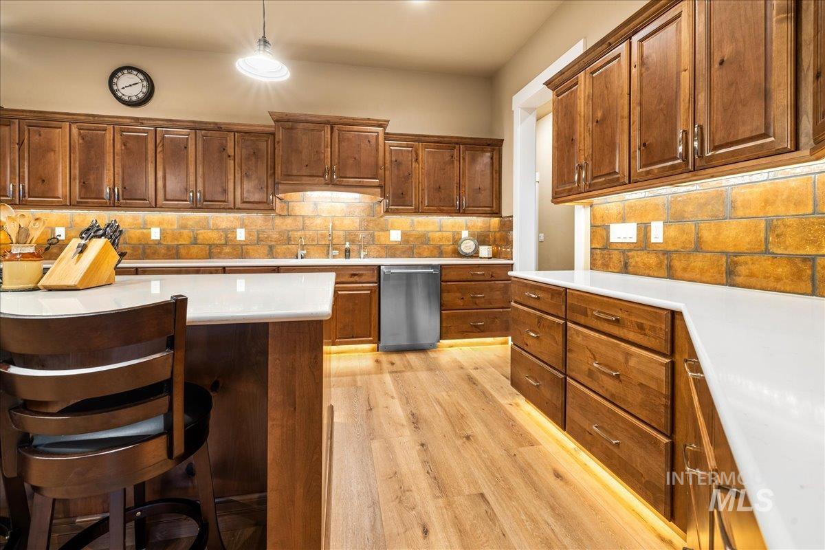Kitchen featuring decorative backsplash, brown cabinetry, stainless steel dishwasher, light wood-style floors, and a breakfast bar area