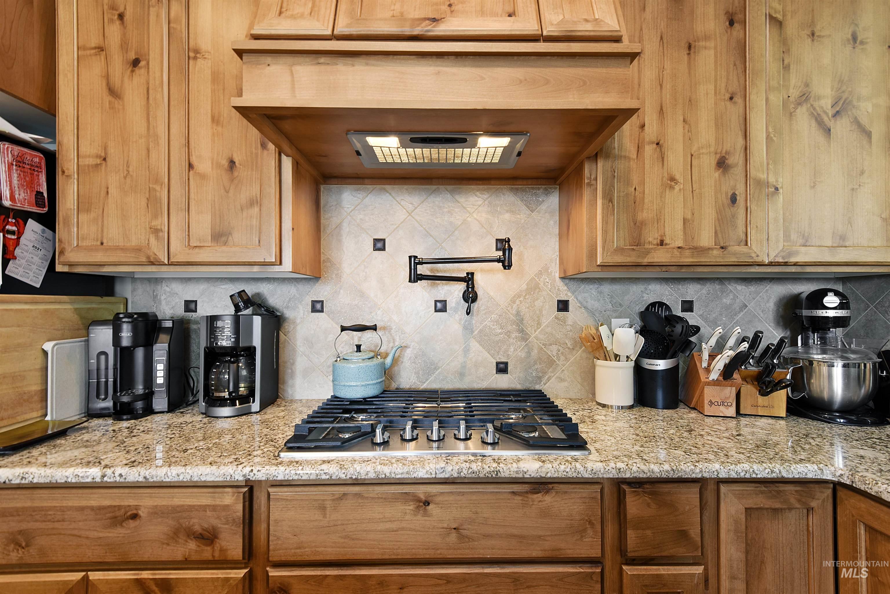 Kitchen featuring decorative backsplash, under cabinet range hood, light stone counters, stainless steel gas cooktop, and brown cabinets