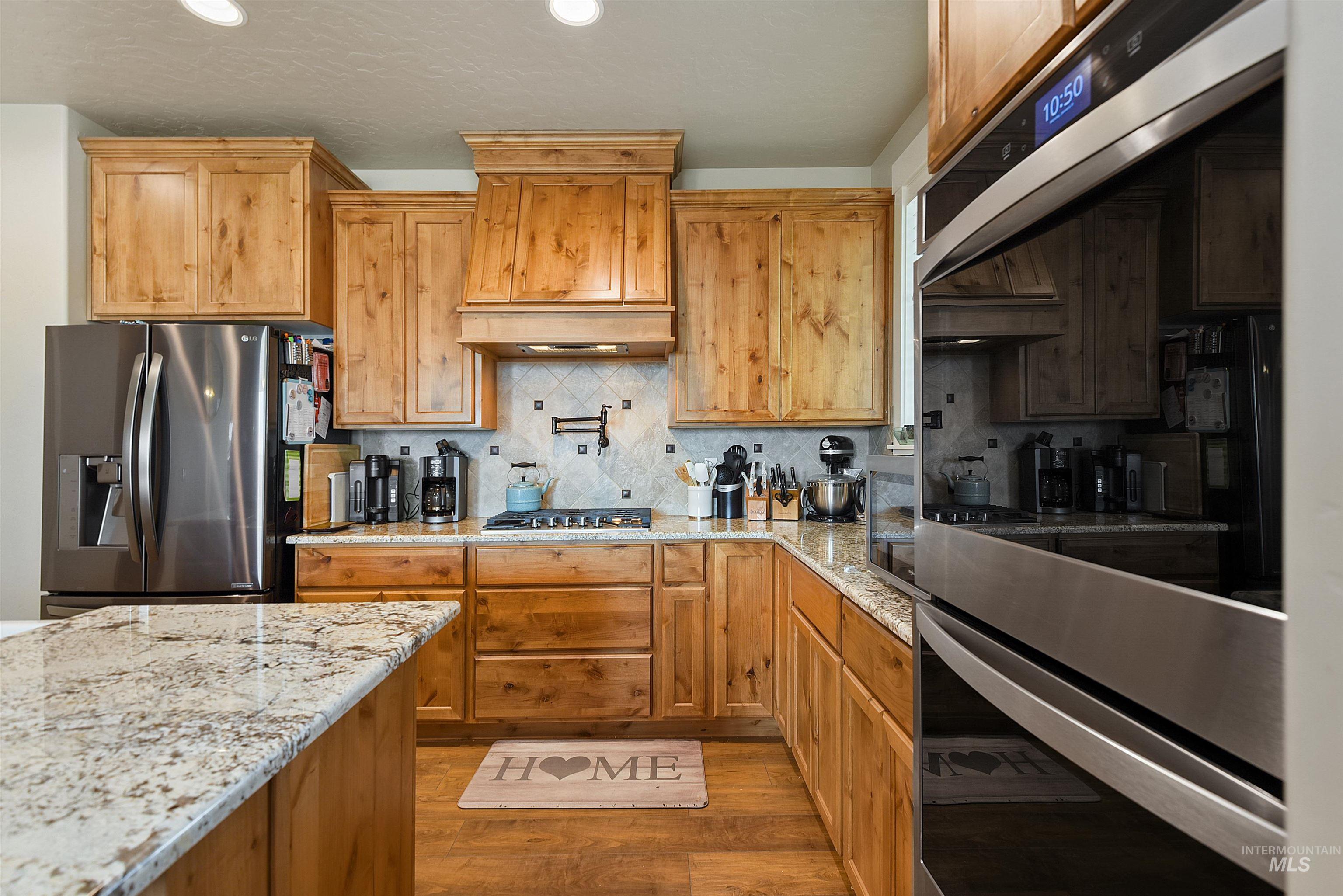 Kitchen featuring tasteful backsplash, appliances with stainless steel finishes, light stone countertops, dark wood finished floors, and brown cabinetry