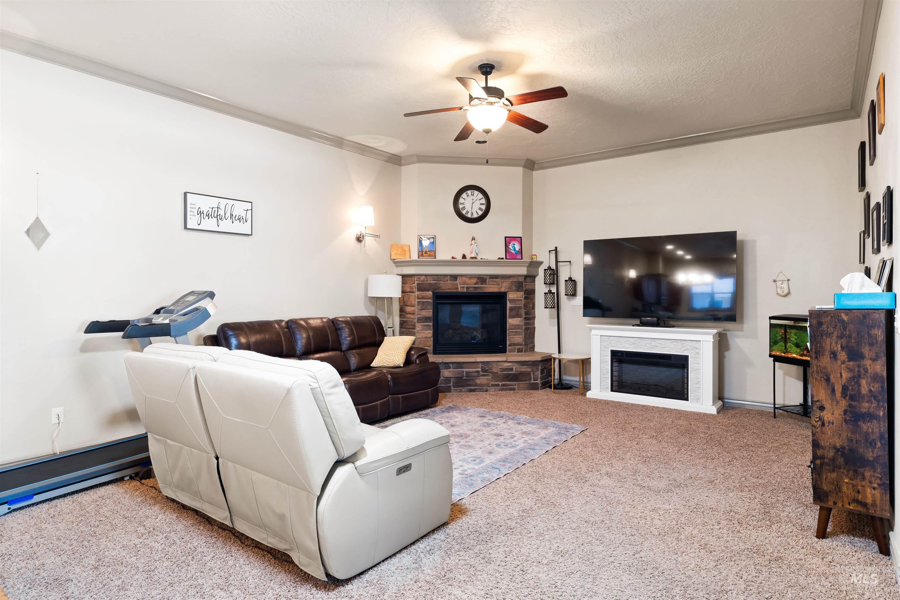 Living room featuring a stone fireplace, carpet flooring, a ceiling fan, crown molding, and a textured ceiling