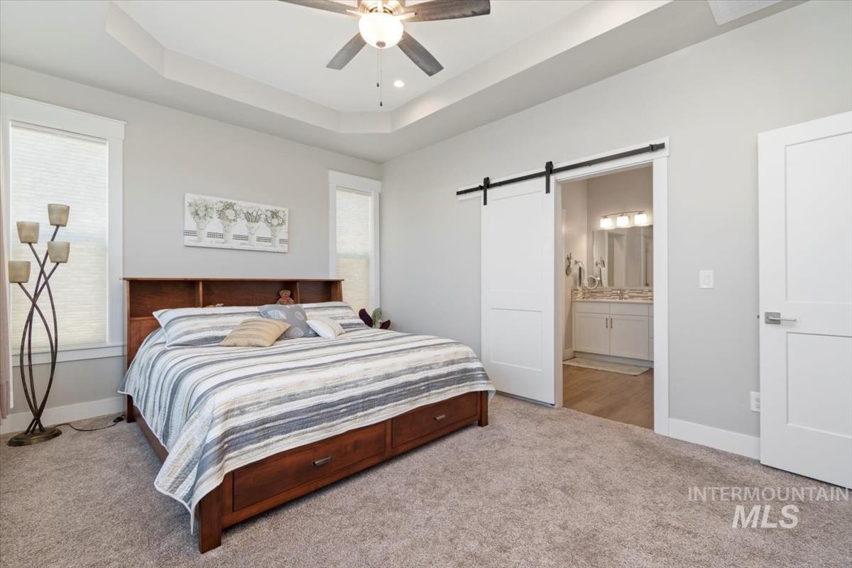 Primary Bedroom featuring a tray ceiling, light carpet, a barn door, a ceiling fan, and ensuite bath