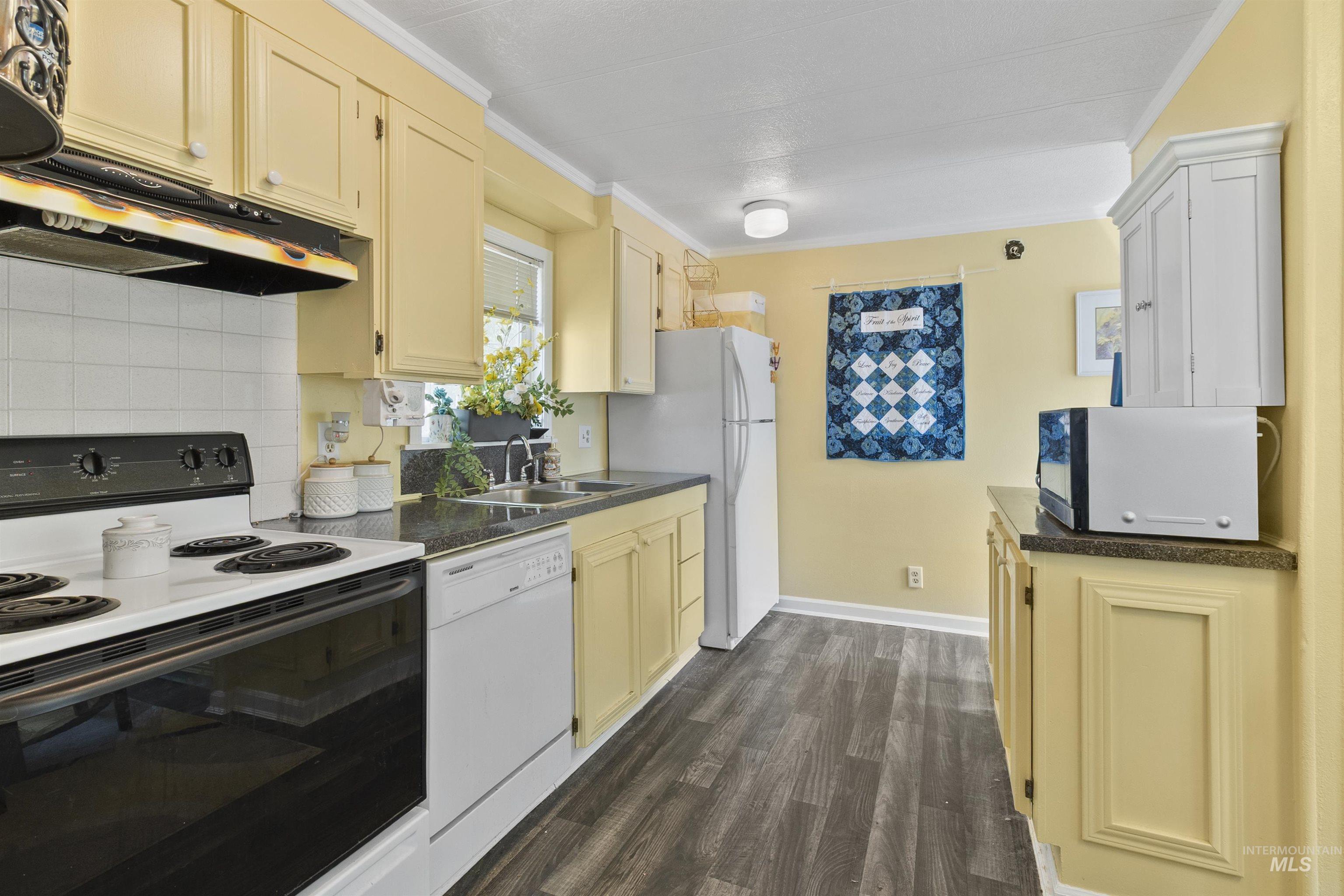 Kitchen featuring white appliances, dark countertops, crown molding, dark wood finished floors, and tasteful backsplash