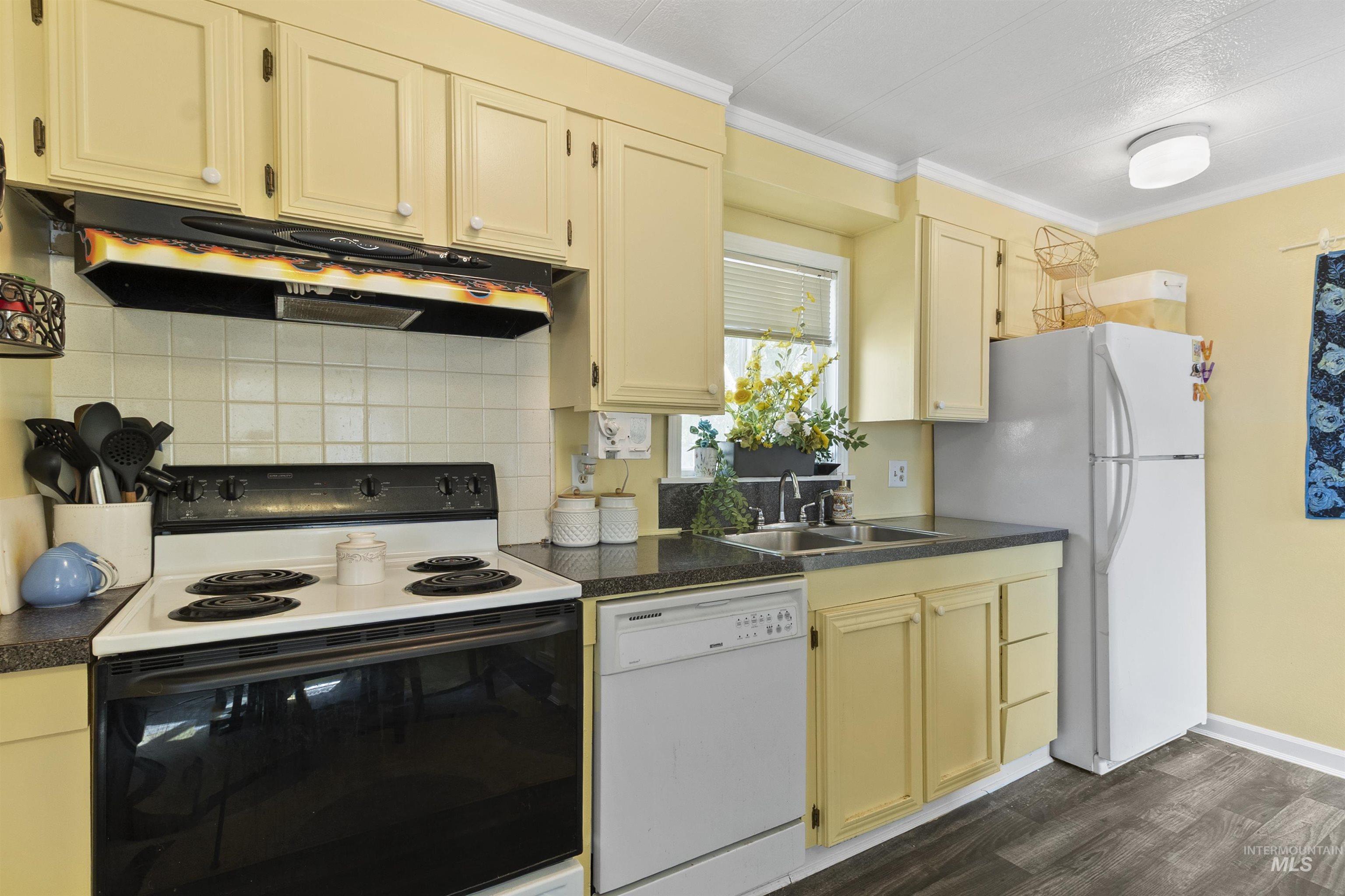 Kitchen featuring white appliances, dark countertops, under cabinet range hood, crown molding, and dark wood finished floors