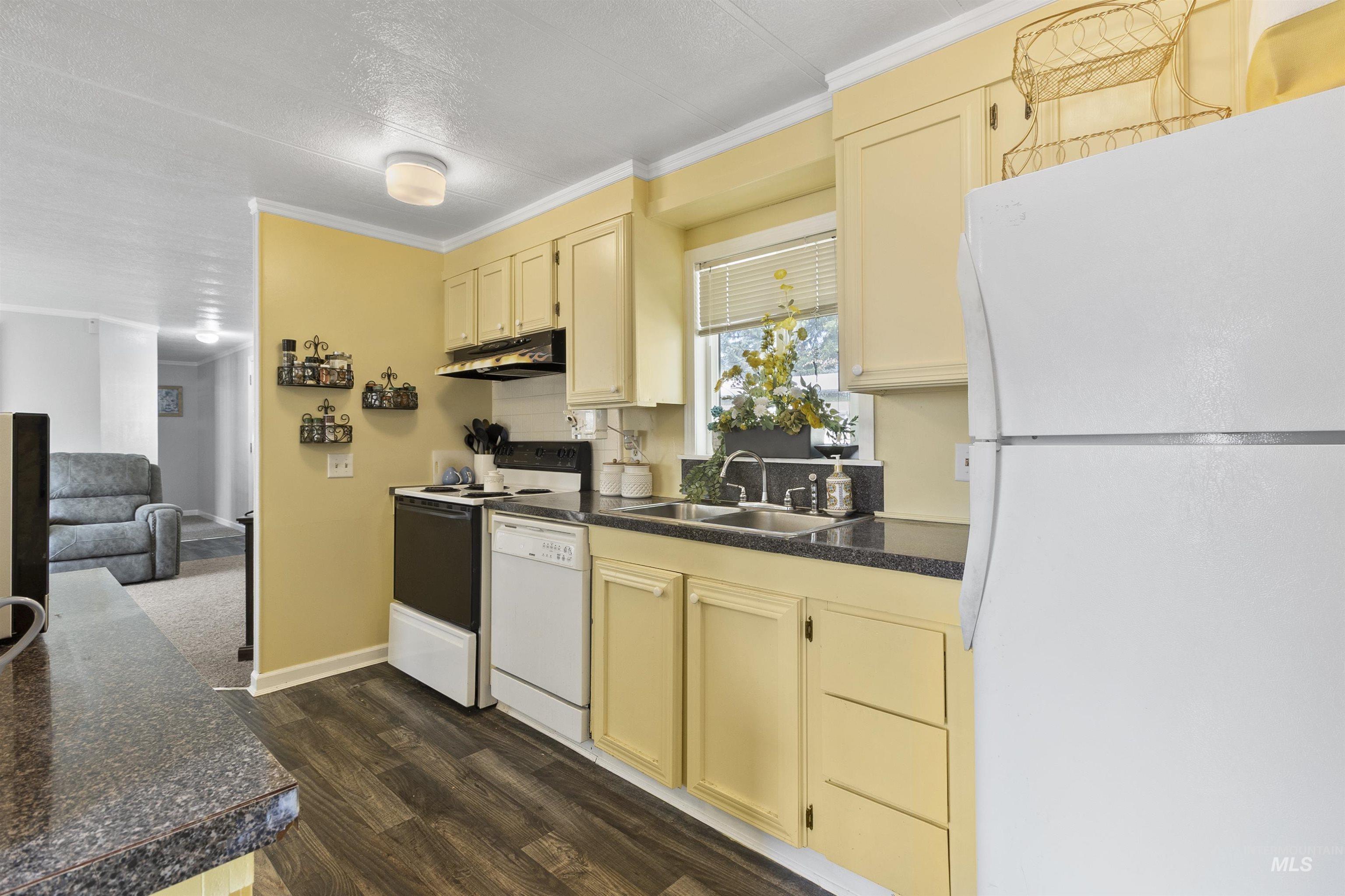 Kitchen with dark countertops, white appliances, crown molding, cream cabinetry, and dark wood-style floors
