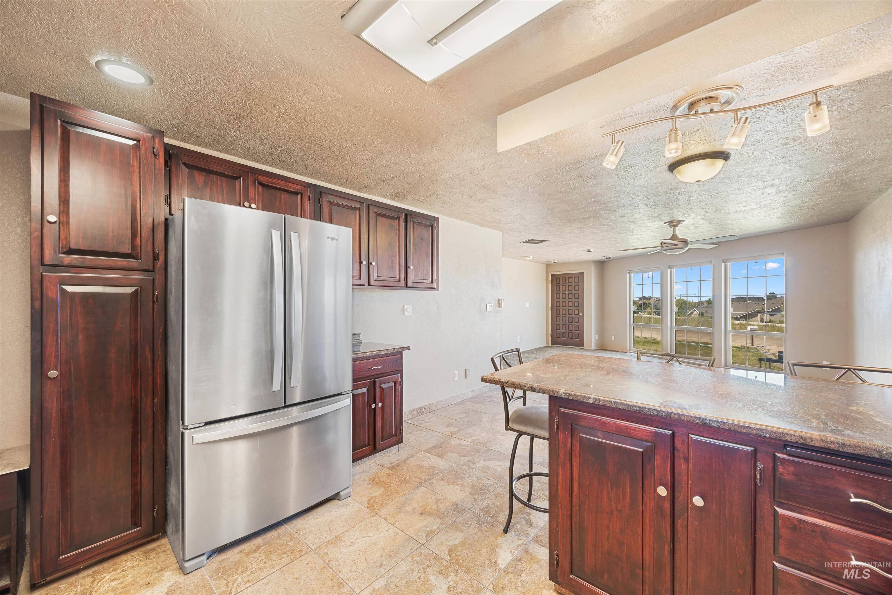 Kitchen with freestanding refrigerator, a textured ceiling, reddish brown cabinets, a kitchen bar, and ceiling fan