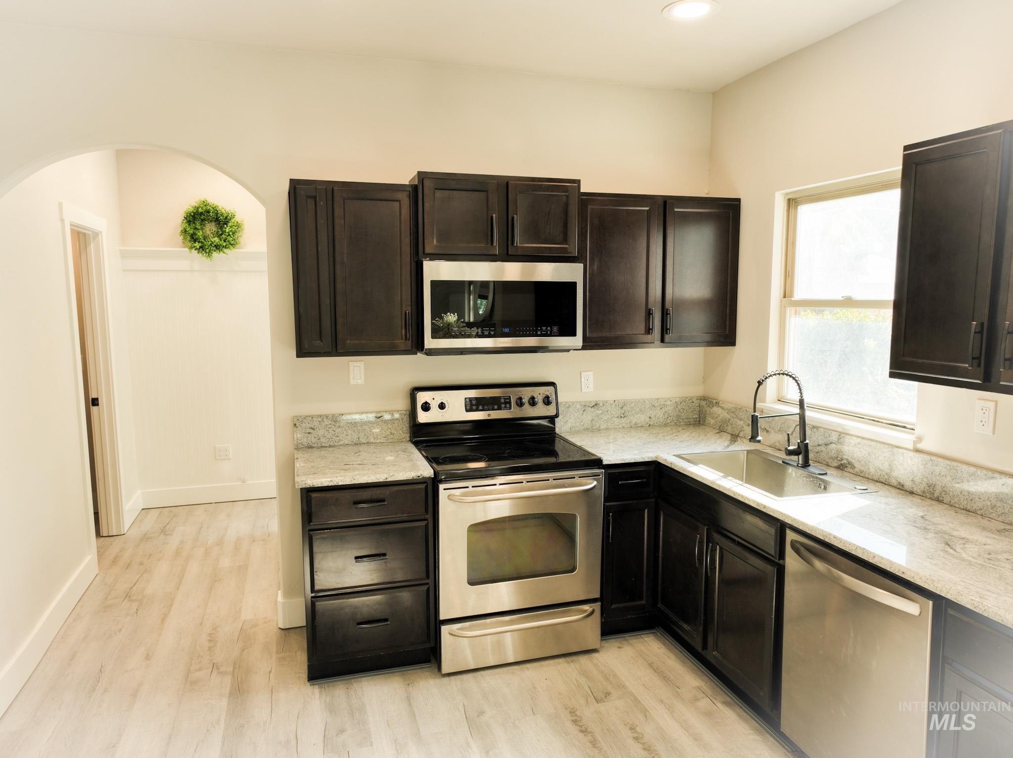 Kitchen with appliances with stainless steel finishes, light stone counters, light wood finished floors, dark brown cabinets, and arched walkways