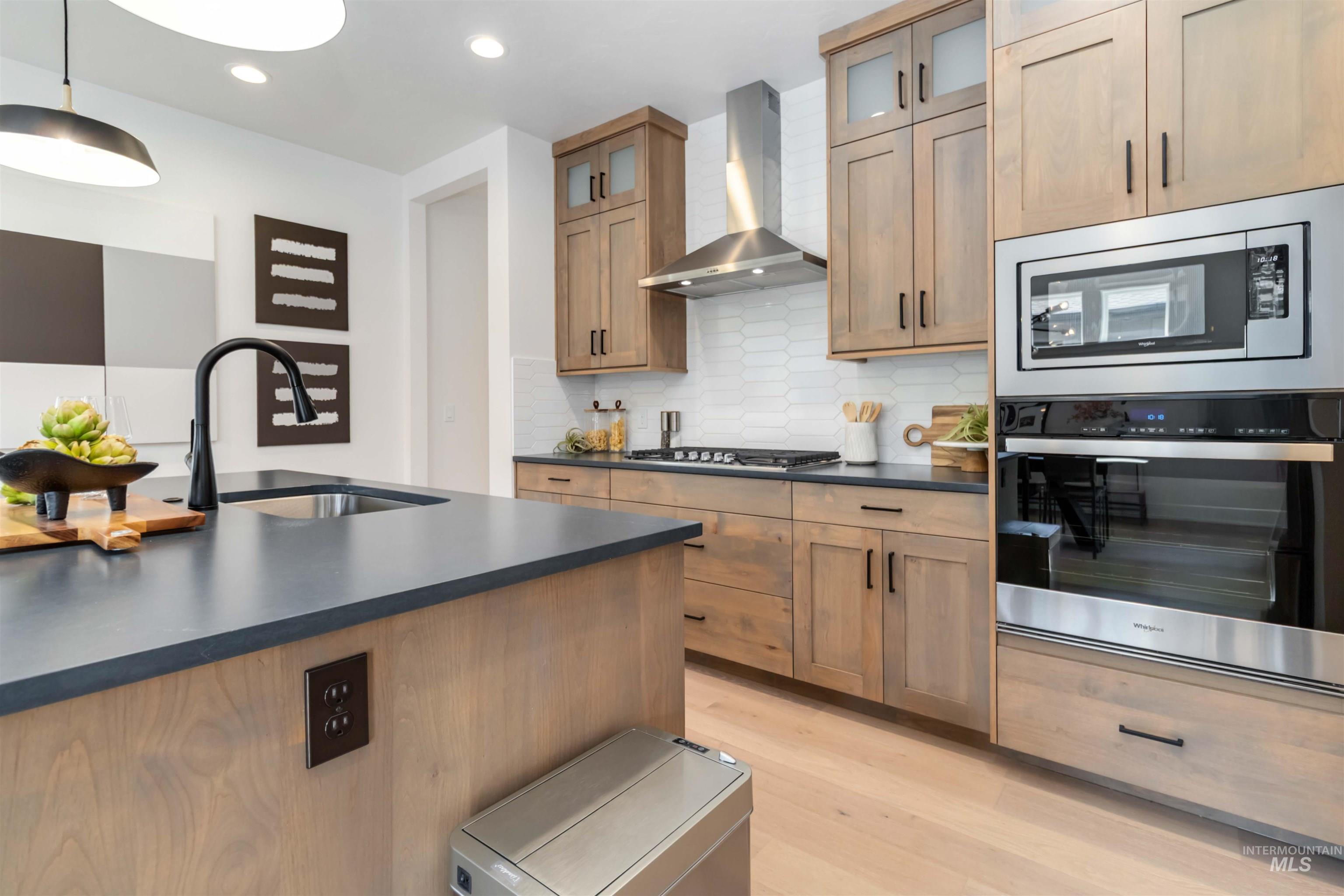 Kitchen featuring tasteful backsplash, stainless steel appliances, wall chimney range hood, glass insert cabinets, and light wood-style floors