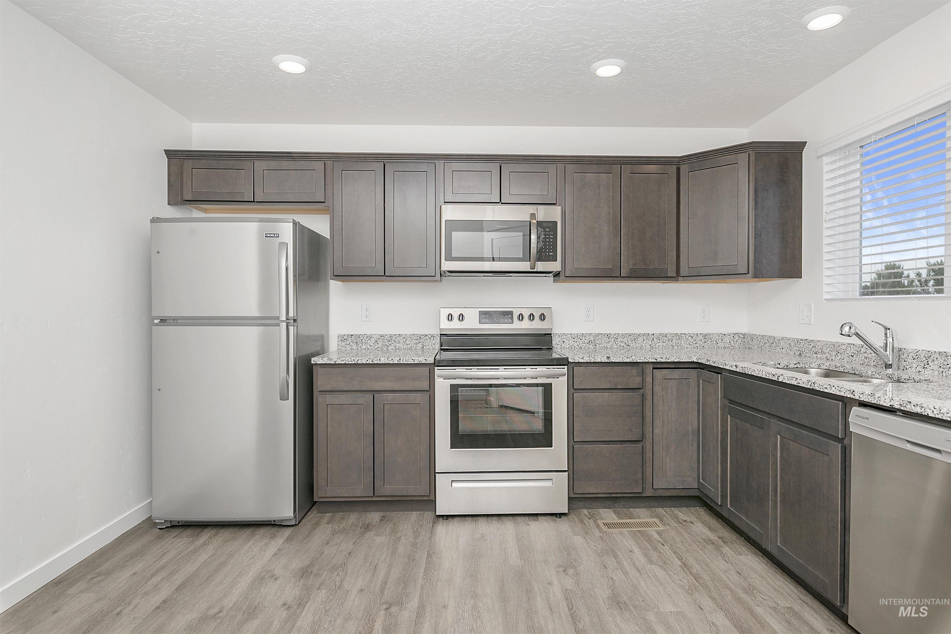 Kitchen featuring appliances with stainless steel finishes, light stone countertops, light wood-style flooring, dark brown cabinets, and a textured ceiling