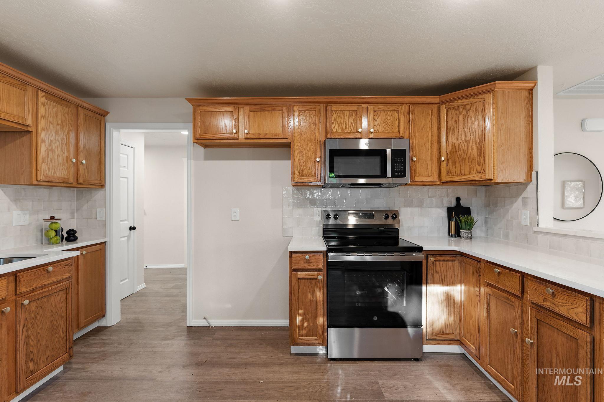 Kitchen with backsplash, appliances with stainless steel finishes, brown cabinetry, light stone countertops, and dark wood-type flooring