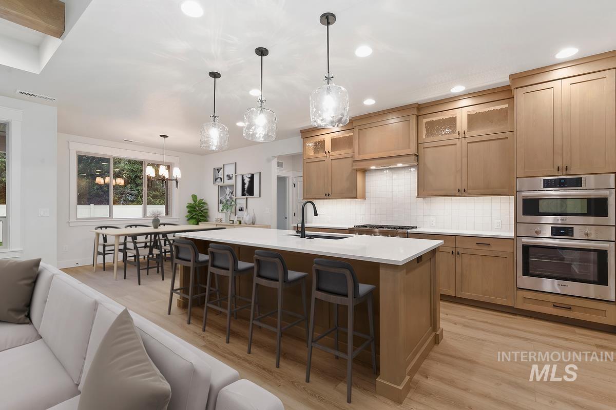 Kitchen with light wood-type flooring, decorative backsplash, light countertops, an island with sink, and a kitchen bar
