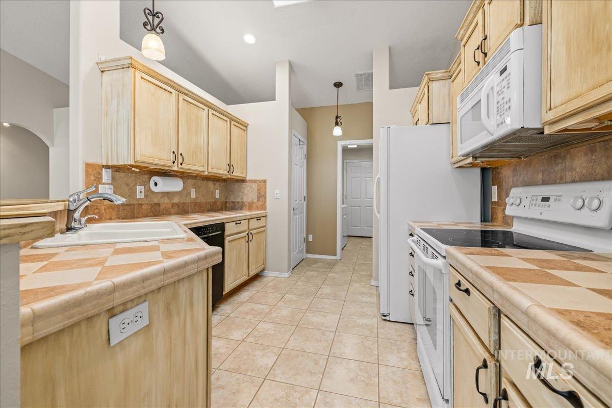 Kitchen with white appliances, tile countertops, light brown cabinets, pendant lighting, and backsplash