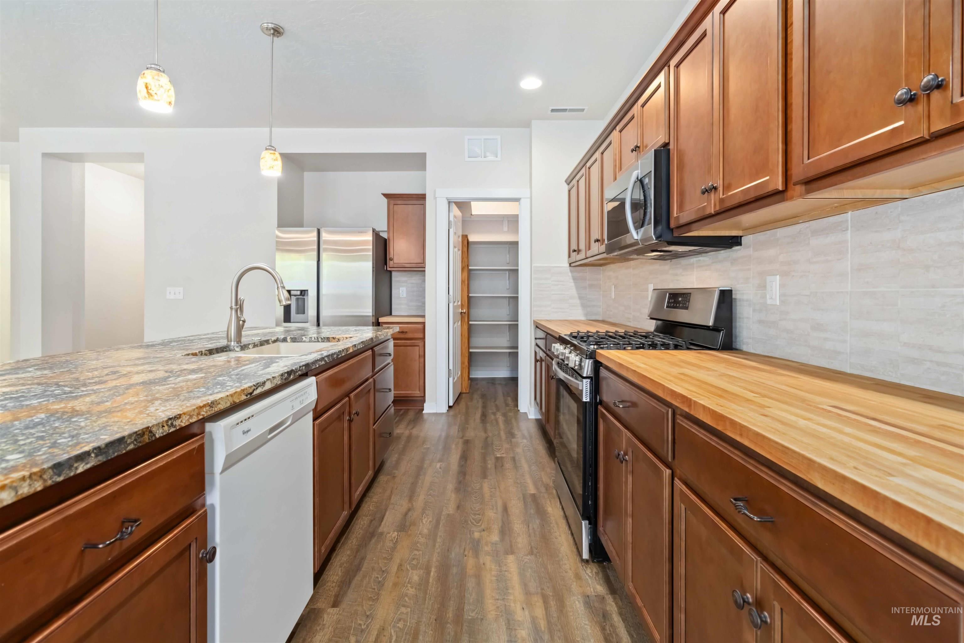 Kitchen with brown cabinets, appliances with stainless steel finishes, dark wood finished floors, backsplash, and hanging light fixtures