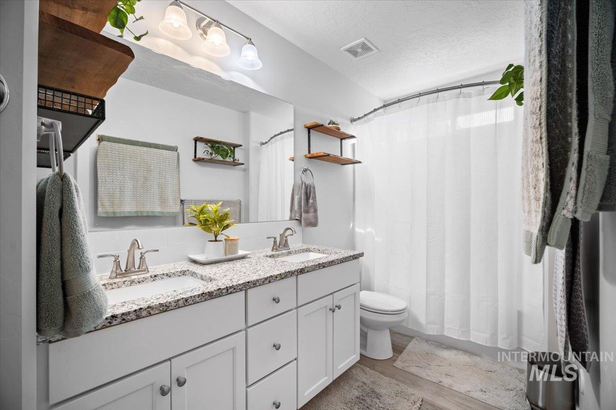 Bathroom with light wood finished floors, a textured ceiling, double vanity, shower / bathtub combination with curtain, and decorative backsplash
