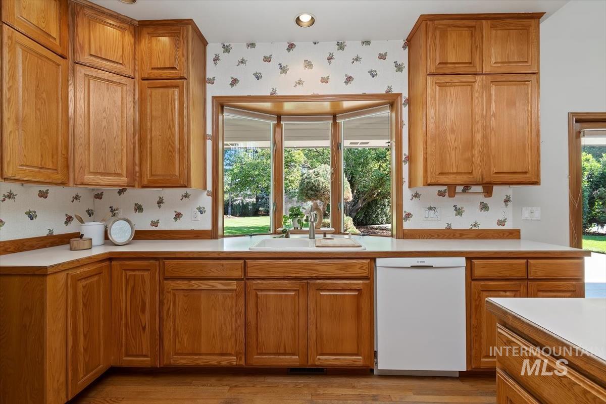 Kitchen featuring wallpapered walls, light countertops, white dishwasher, brown cabinets, and plenty of natural light