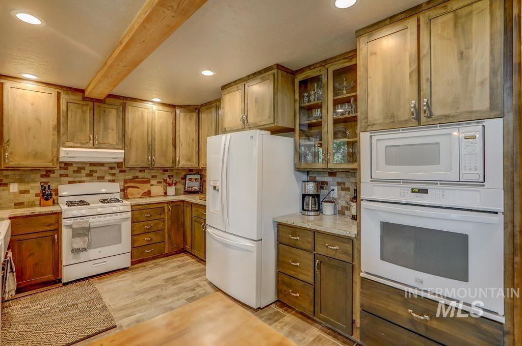 Kitchen featuring white appliances, tasteful backsplash, glass insert cabinets, brown cabinetry, and beamed ceiling