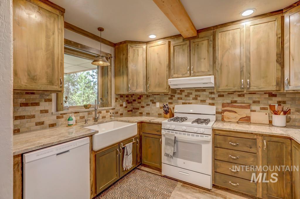 Kitchen featuring white appliances, tasteful backsplash, hanging light fixtures, under cabinet range hood, and light stone counters