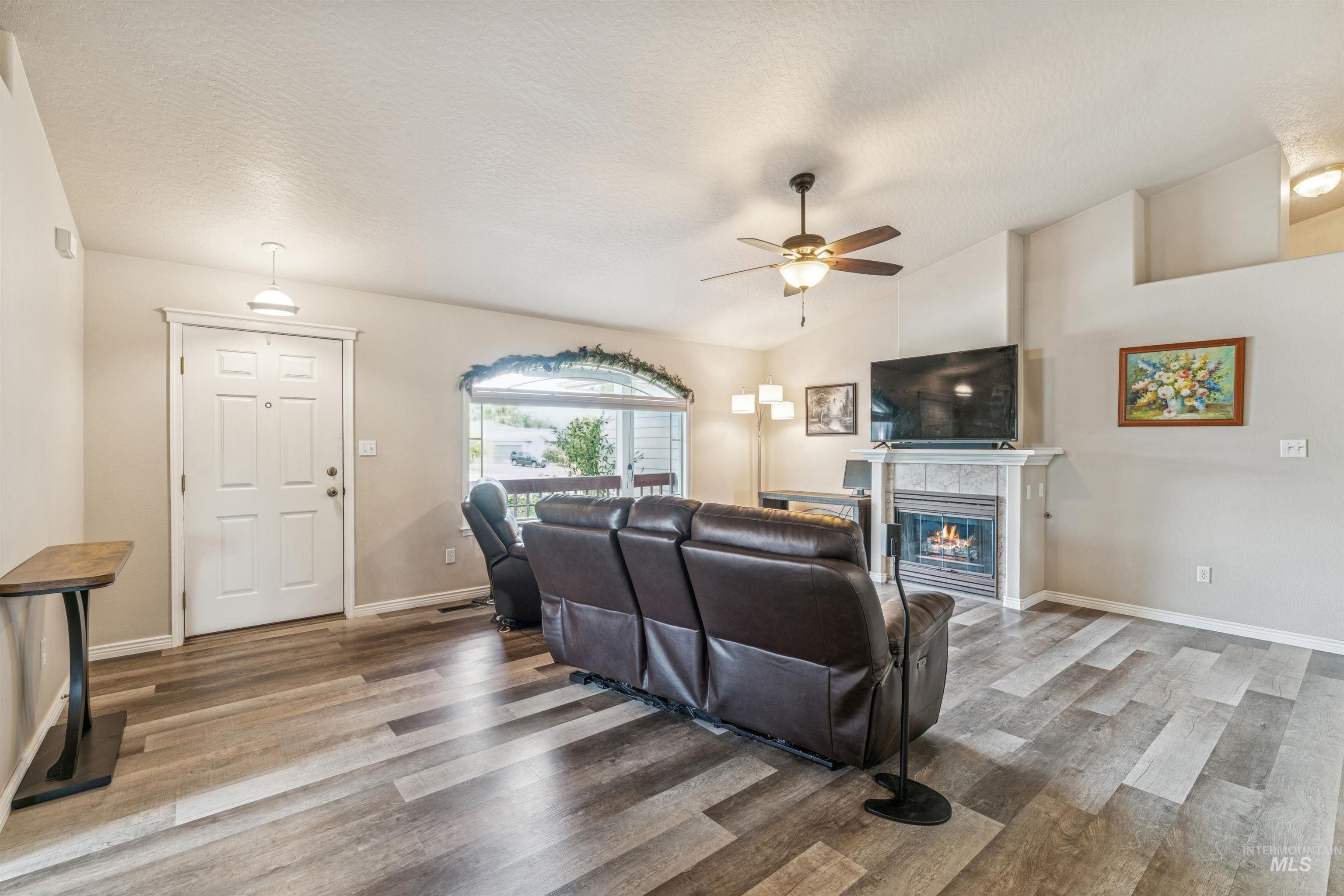 Living room with light wood finished floors, a tiled fireplace, vaulted ceiling, a ceiling fan, and a textured ceiling