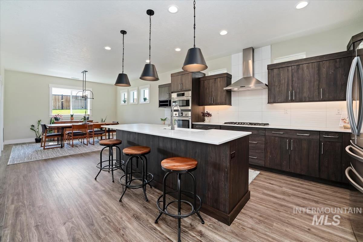 Kitchen with backsplash, dark brown cabinetry, hanging light fixtures, wall chimney exhaust hood, and a center island with sink