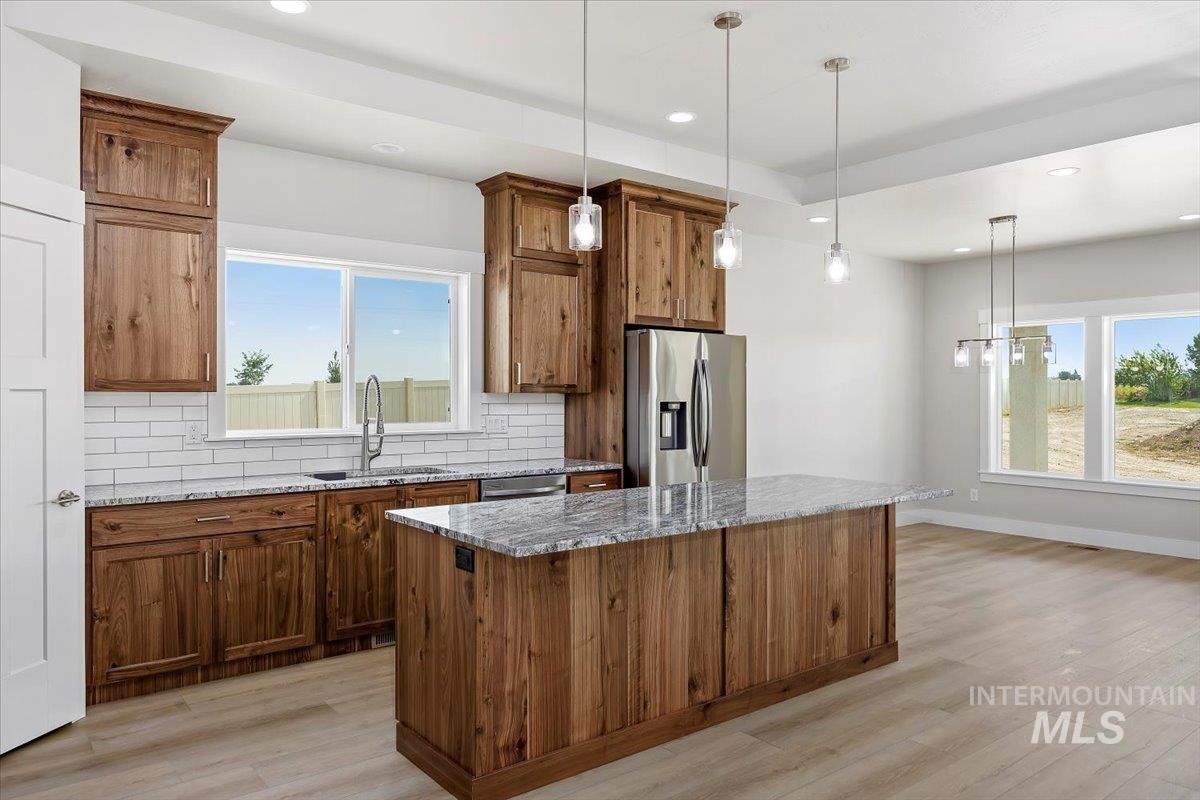 Kitchen with light stone counters, pendant lighting, brown cabinetry, tasteful backsplash, and stainless steel fridge with ice dispenser