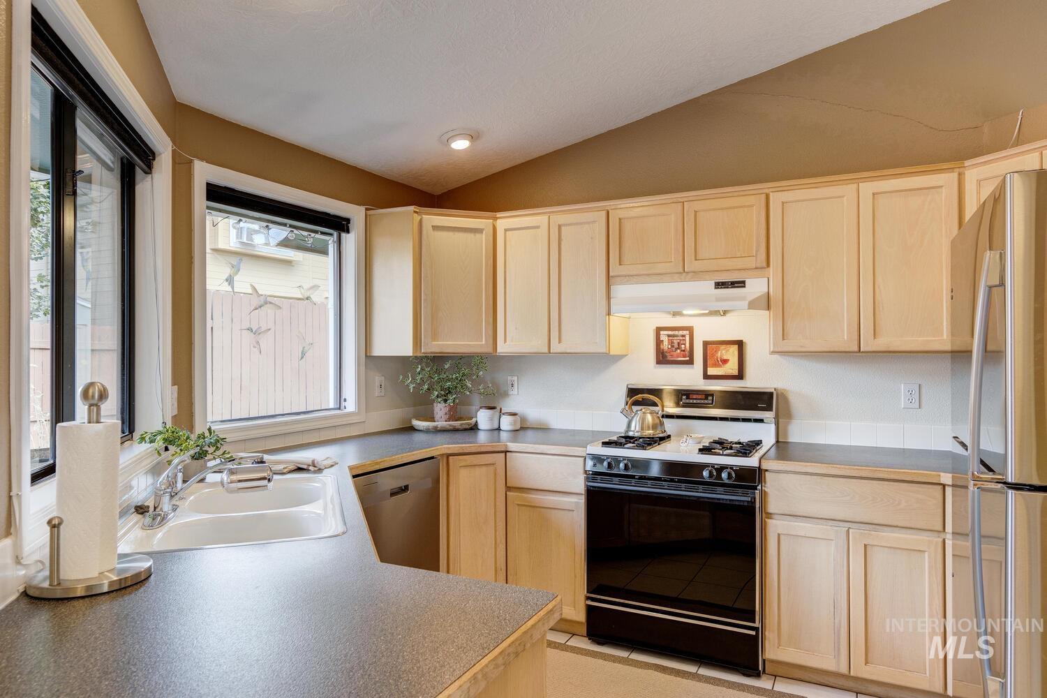 Kitchen featuring light brown cabinetry, stainless steel appliances, vaulted ceiling, under cabinet range hood, and light tile patterned floors