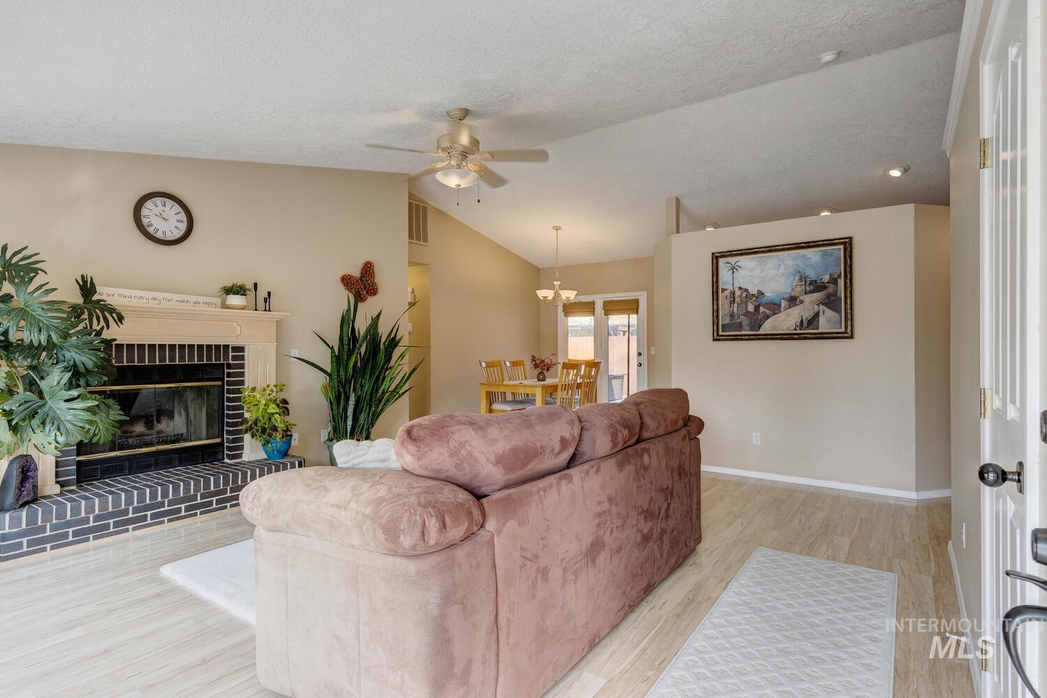 Living room featuring light wood-style flooring, ceiling fan, vaulted ceiling, a brick fireplace, and a chandelier