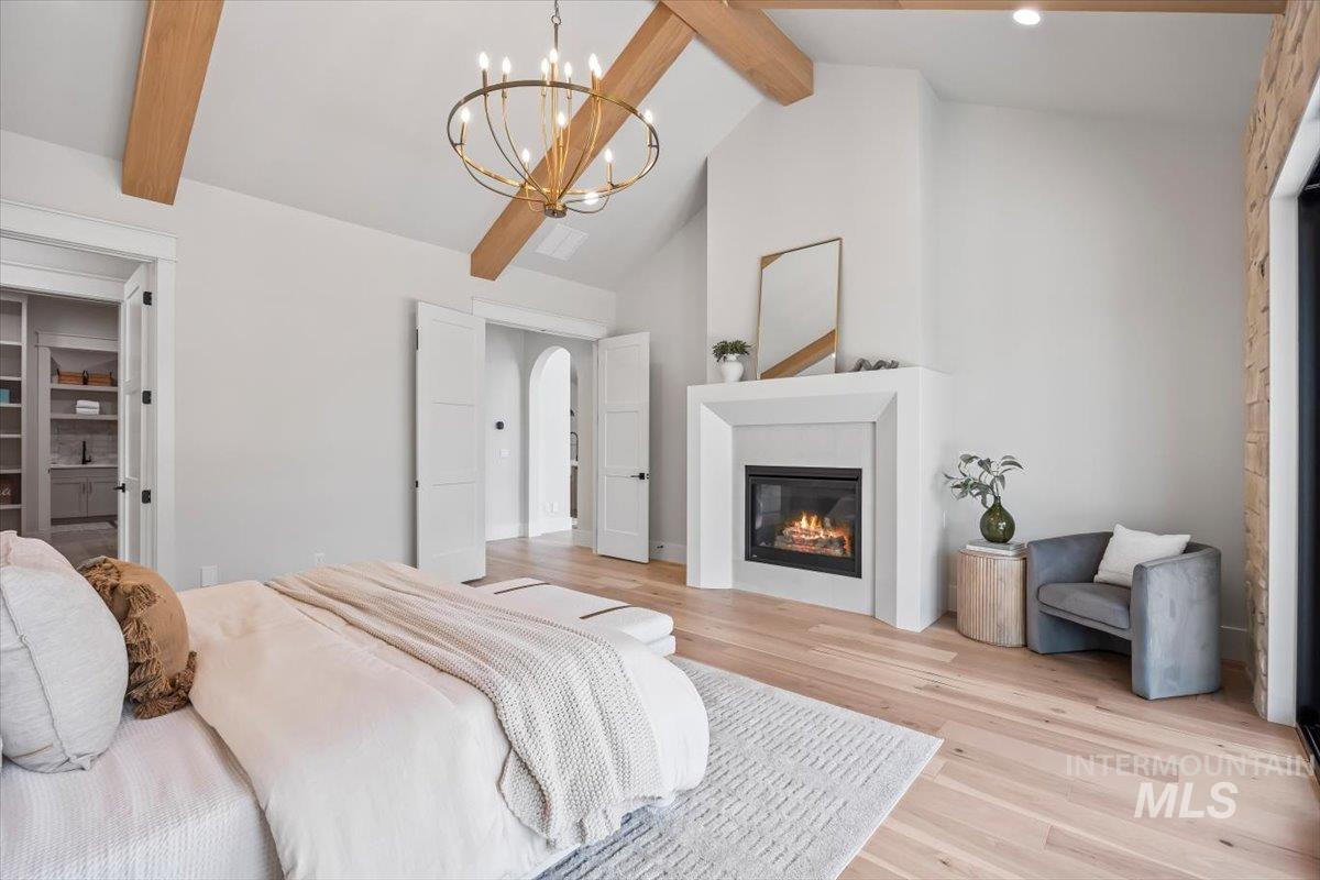 Bedroom with beamed ceiling, light wood finished floors, high vaulted ceiling, arched walkways, and a glass covered fireplace