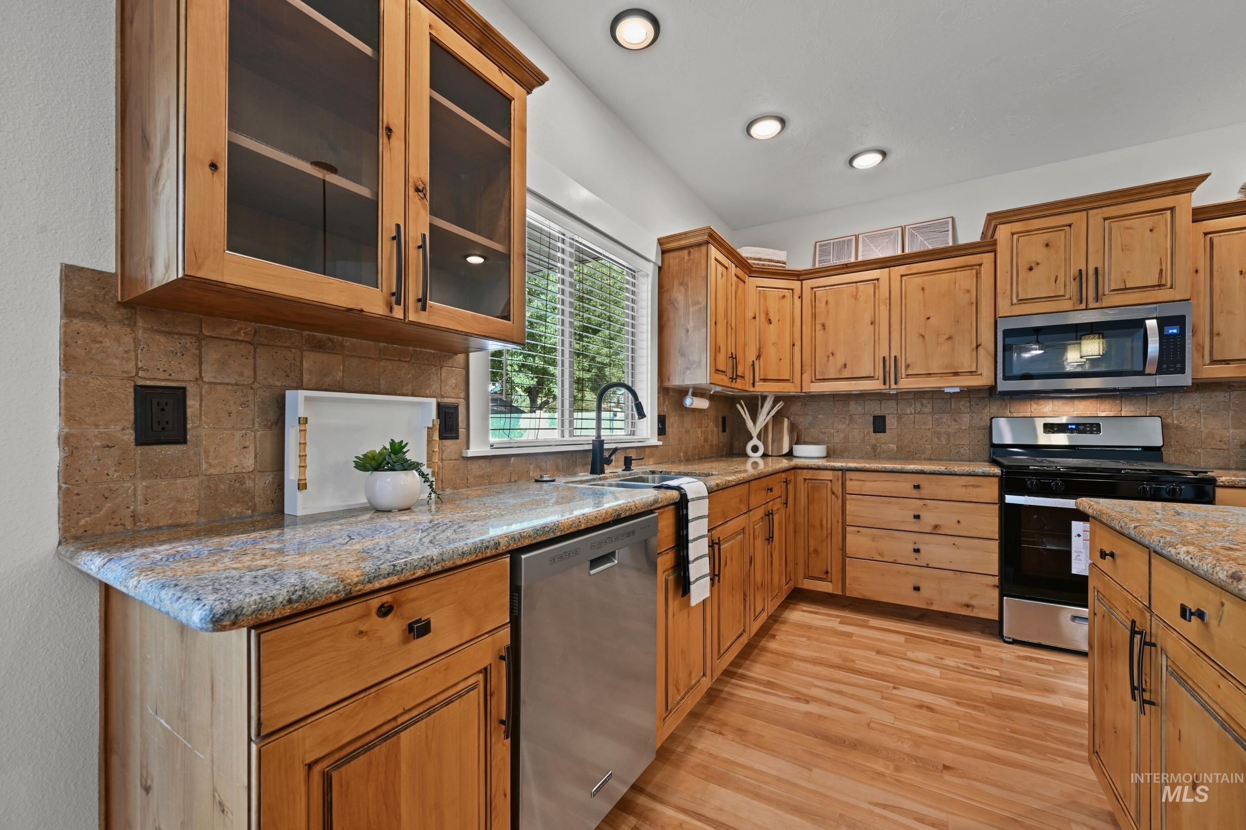 Kitchen with appliances with stainless steel finishes, backsplash, glass insert cabinets, light stone counters, and light wood-style floors