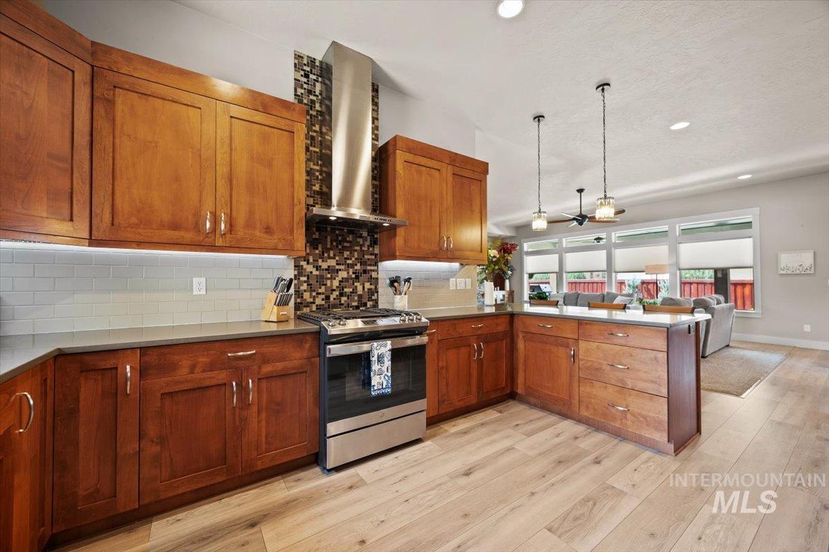 Kitchen with backsplash, brown cabinetry, stainless steel gas stove, wall chimney exhaust hood, and a peninsula
