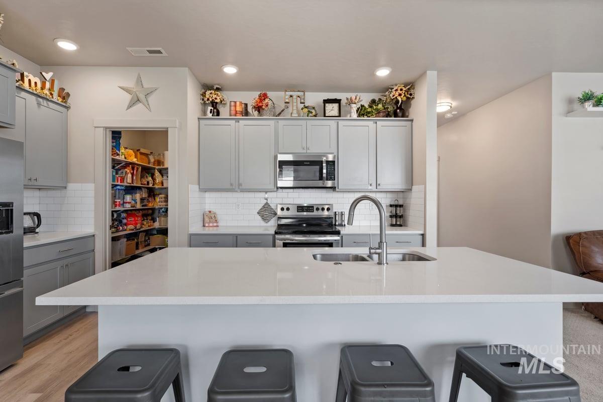 Kitchen featuring gray cabinets, appliances with stainless steel finishes, tasteful backsplash, a kitchen island with sink, and light stone counters