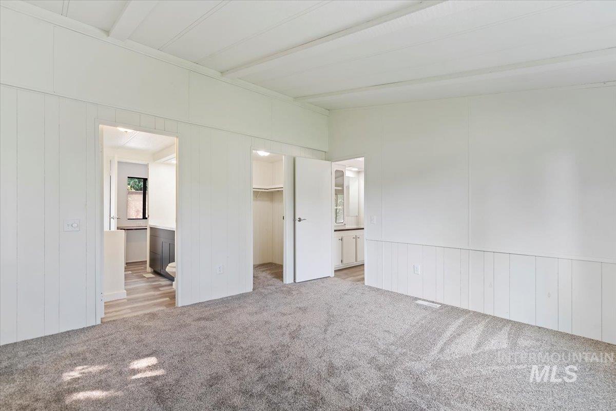 Unfurnished bedroom with ensuite bath, light carpet, a spacious closet, beamed ceiling, and wood walls
