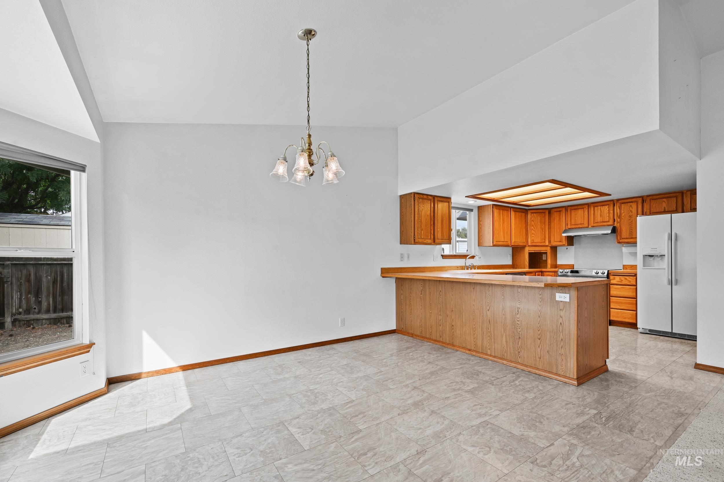 Kitchen featuring a peninsula, light countertops, white fridge with ice dispenser, brown cabinetry, and hanging light fixtures