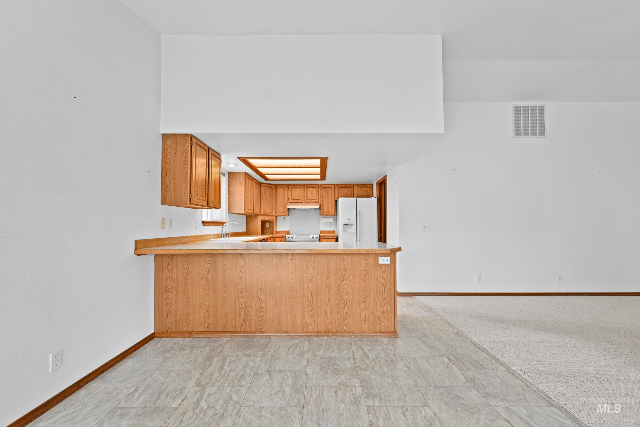 Kitchen with light countertops, a peninsula, white fridge with ice dispenser, and brown cabinets