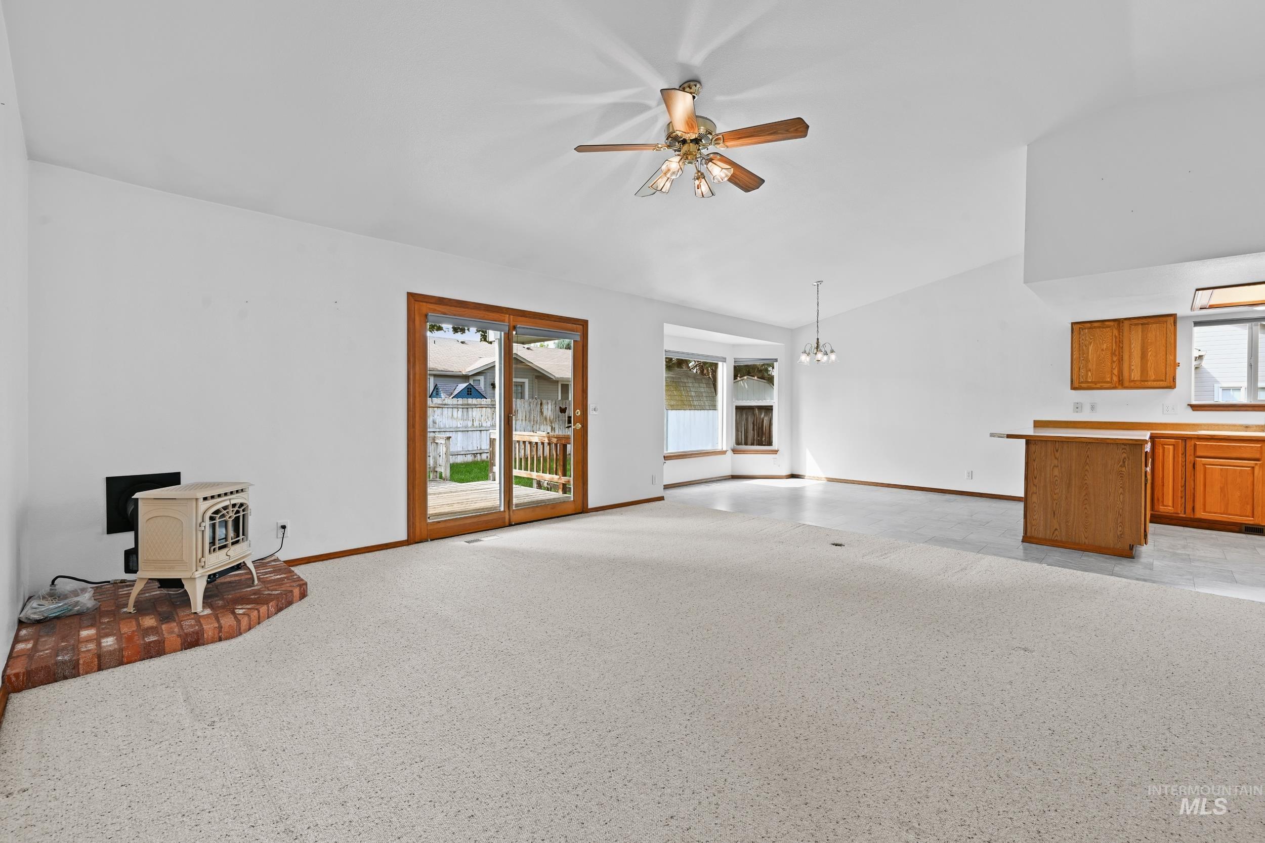 Unfurnished living room with a wood stove, a ceiling fan, a chandelier, vaulted ceiling, and light colored carpet