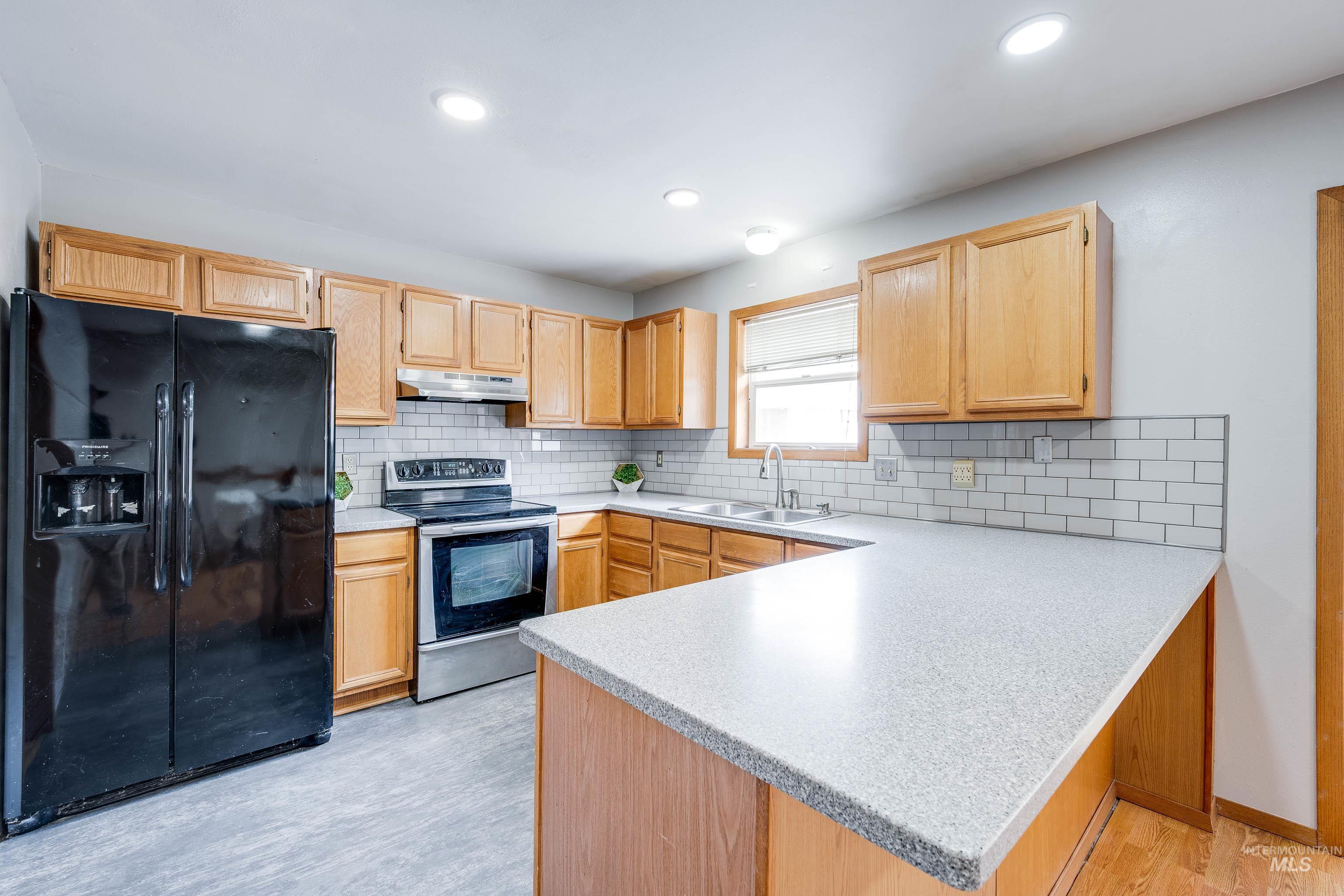 Kitchen with black refrigerator with ice dispenser, stainless steel range with electric stovetop, a peninsula, decorative backsplash, and light wood-style flooring