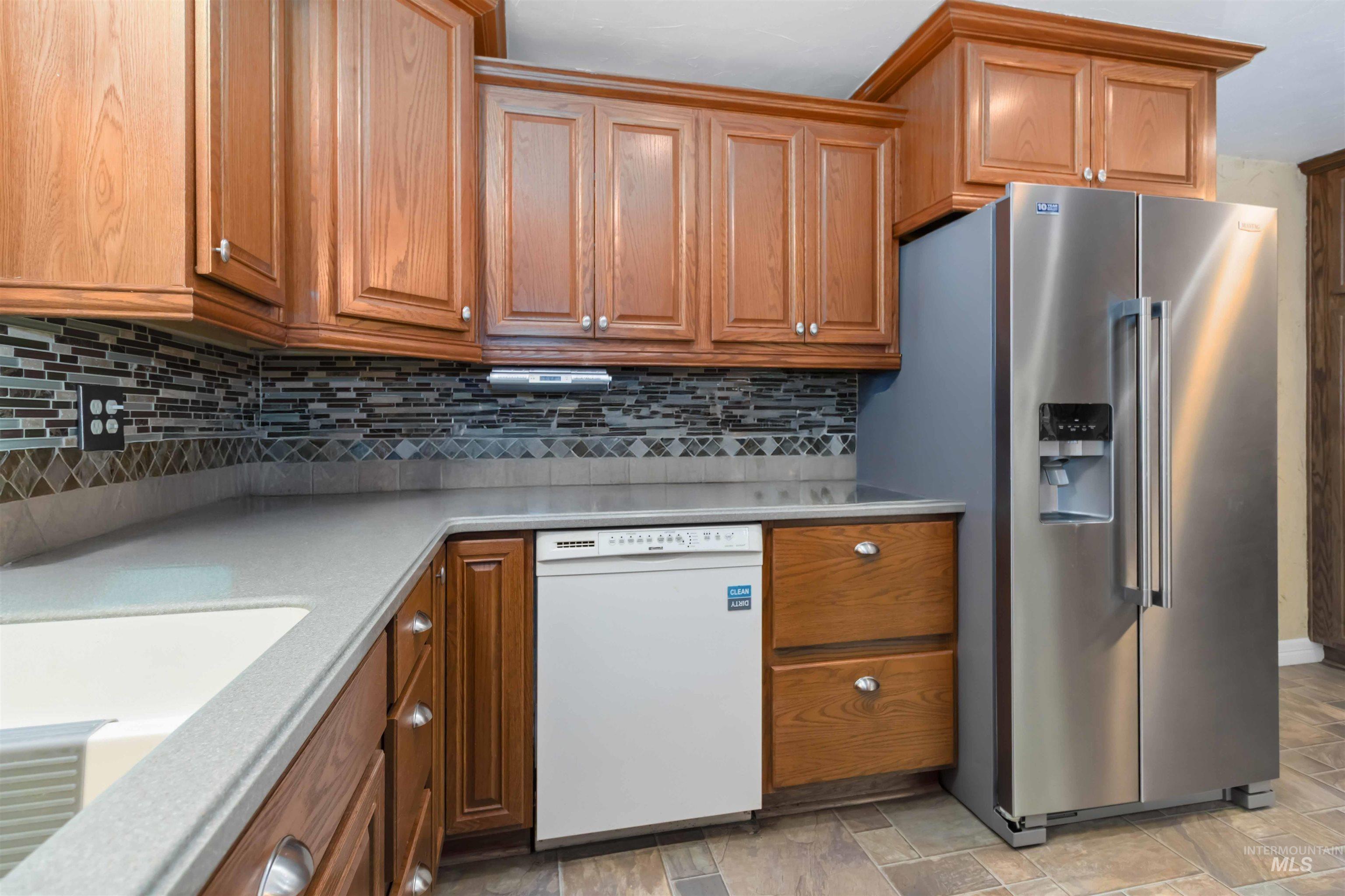 Kitchen featuring brown cabinetry, stainless steel refrigerator with ice dispenser, dishwasher, decorative backsplash, and light stone countertops