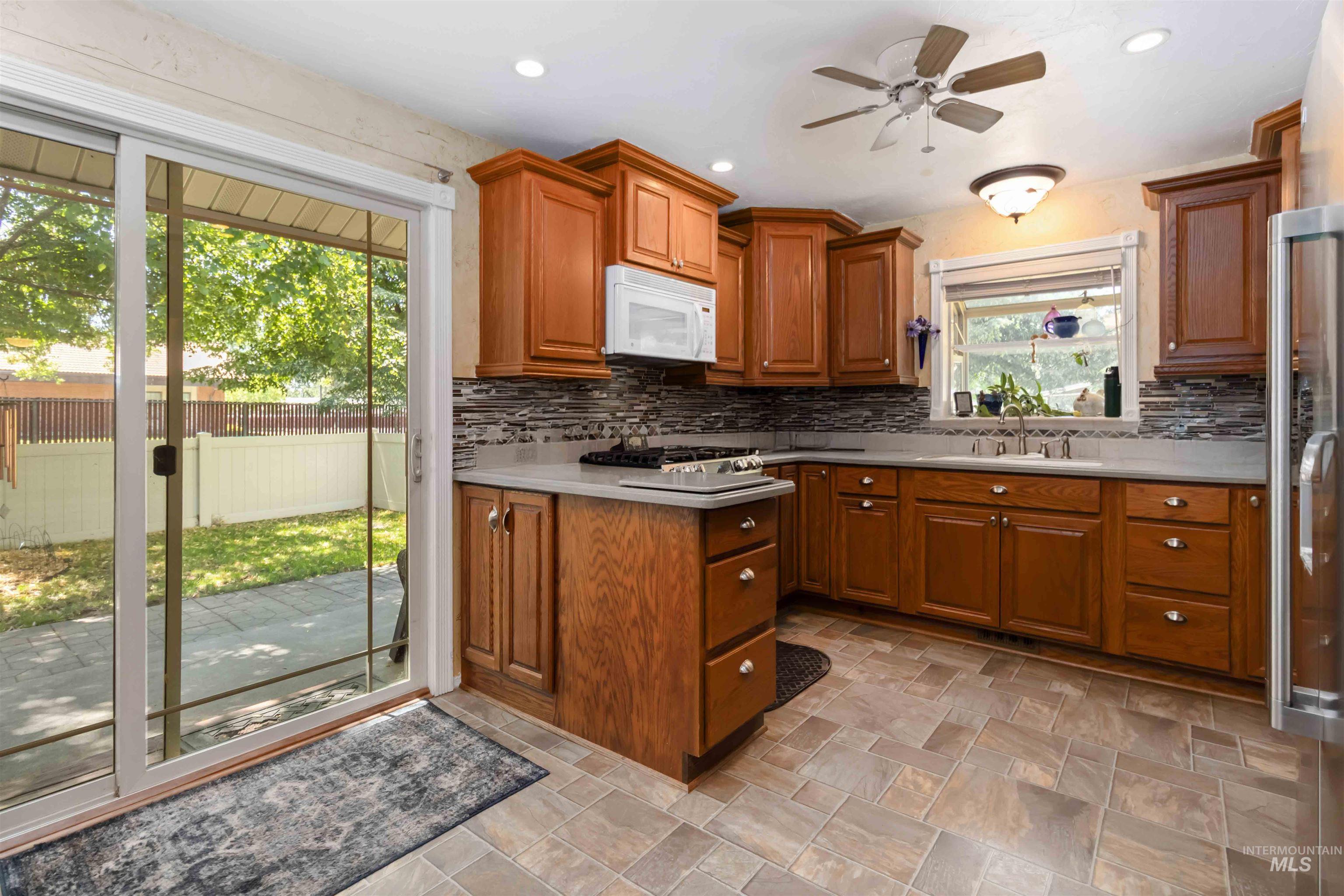Kitchen with stone finish floors, brown cabinetry, backsplash, white microwave, and stainless steel fridge