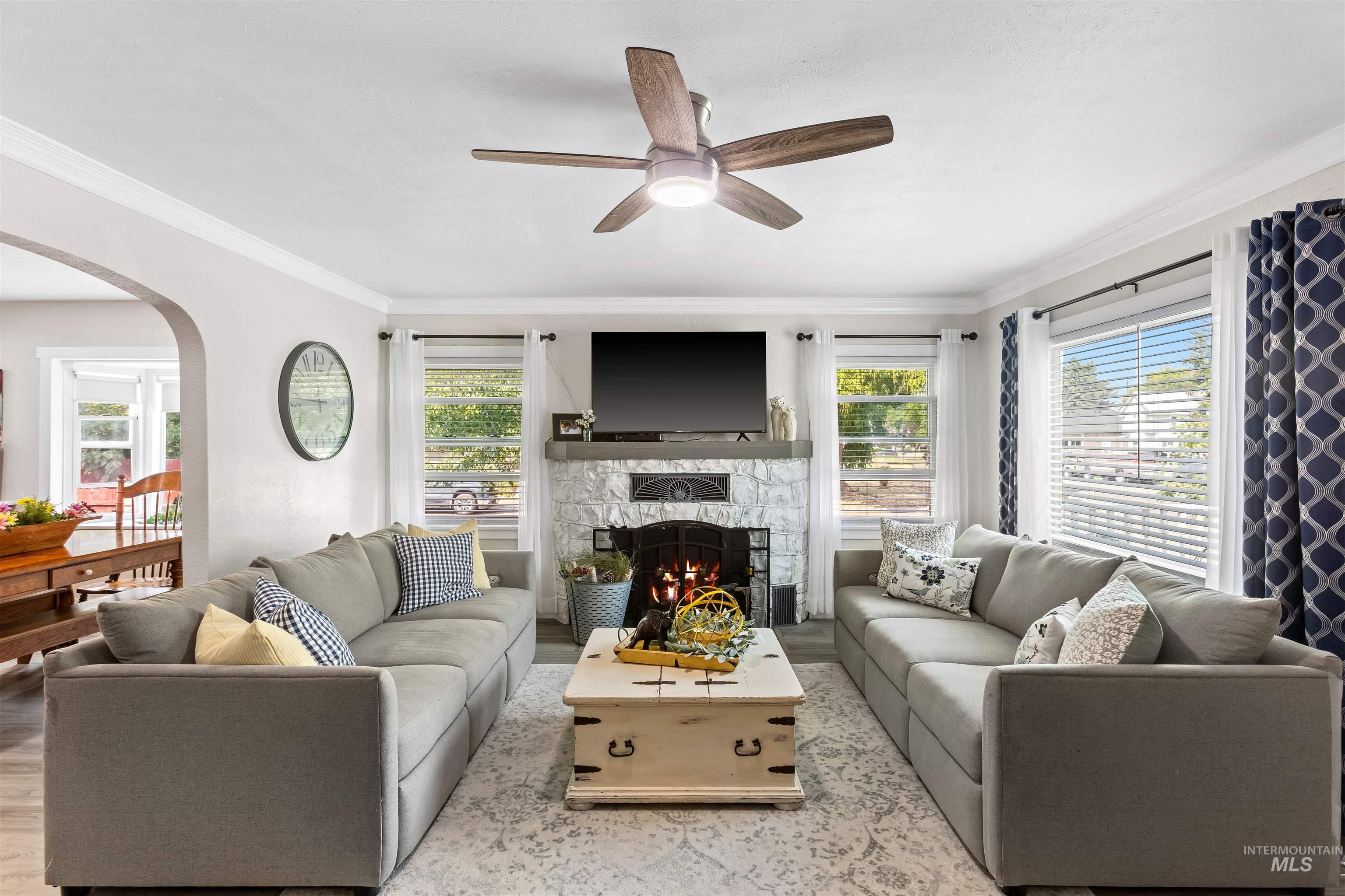 Living area with crown molding, arched walkways, a stone fireplace, wood finished floors, and a ceiling fan