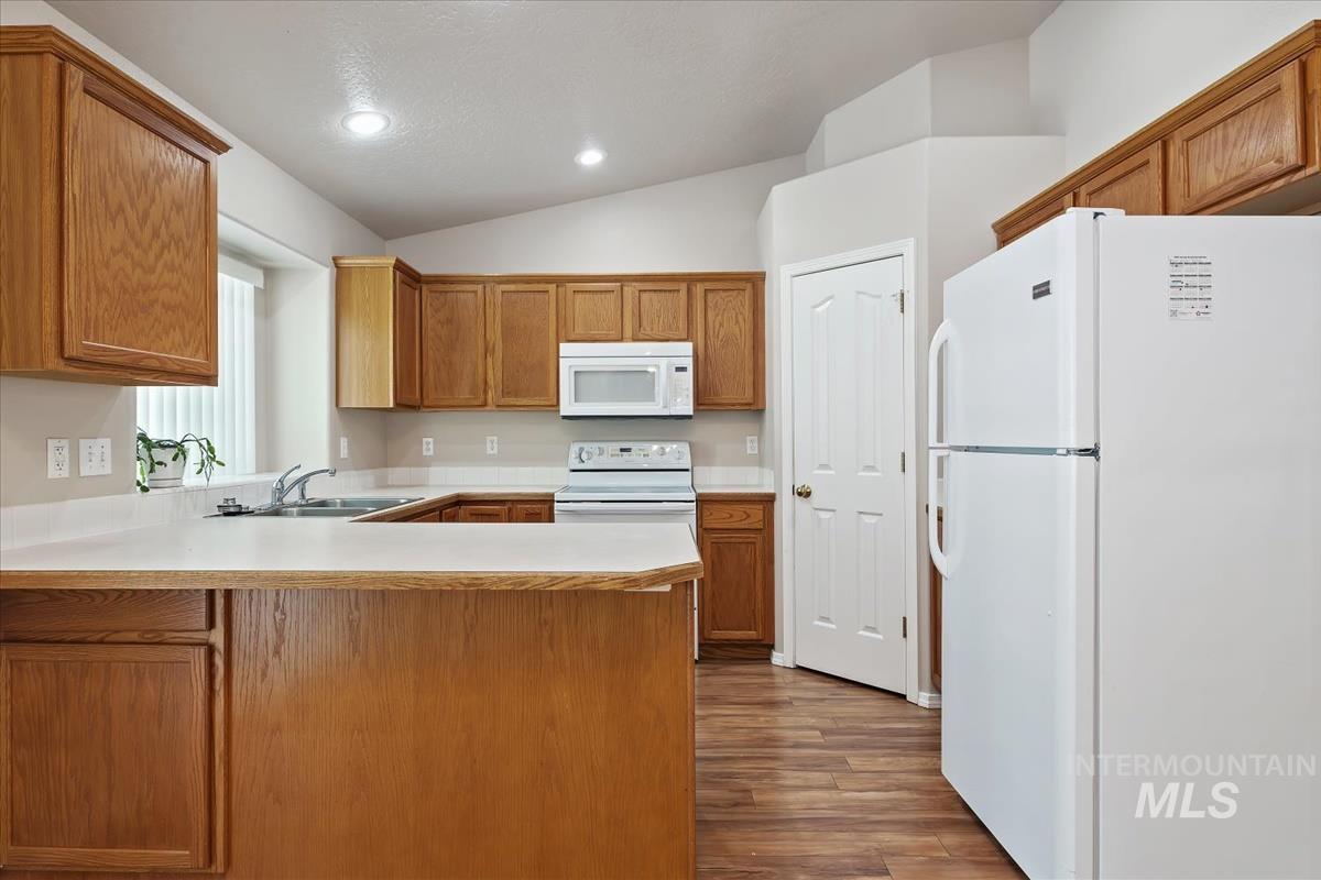 Kitchen featuring white appliances, light countertops, a peninsula, vaulted ceiling, and brown cabinetry