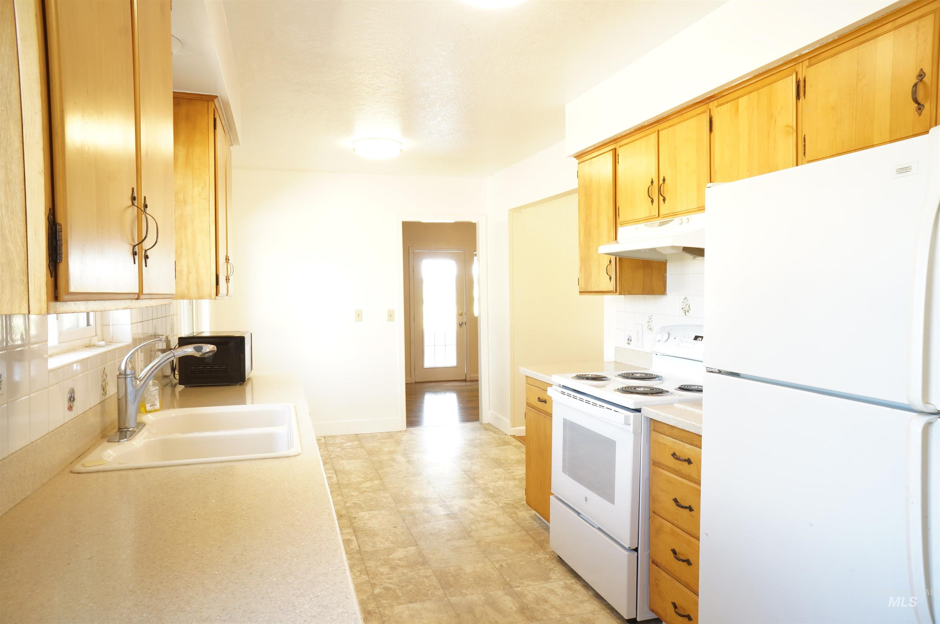 Kitchen featuring white appliances, decorative backsplash, light countertops, under cabinet range hood, and light brown cabinetry