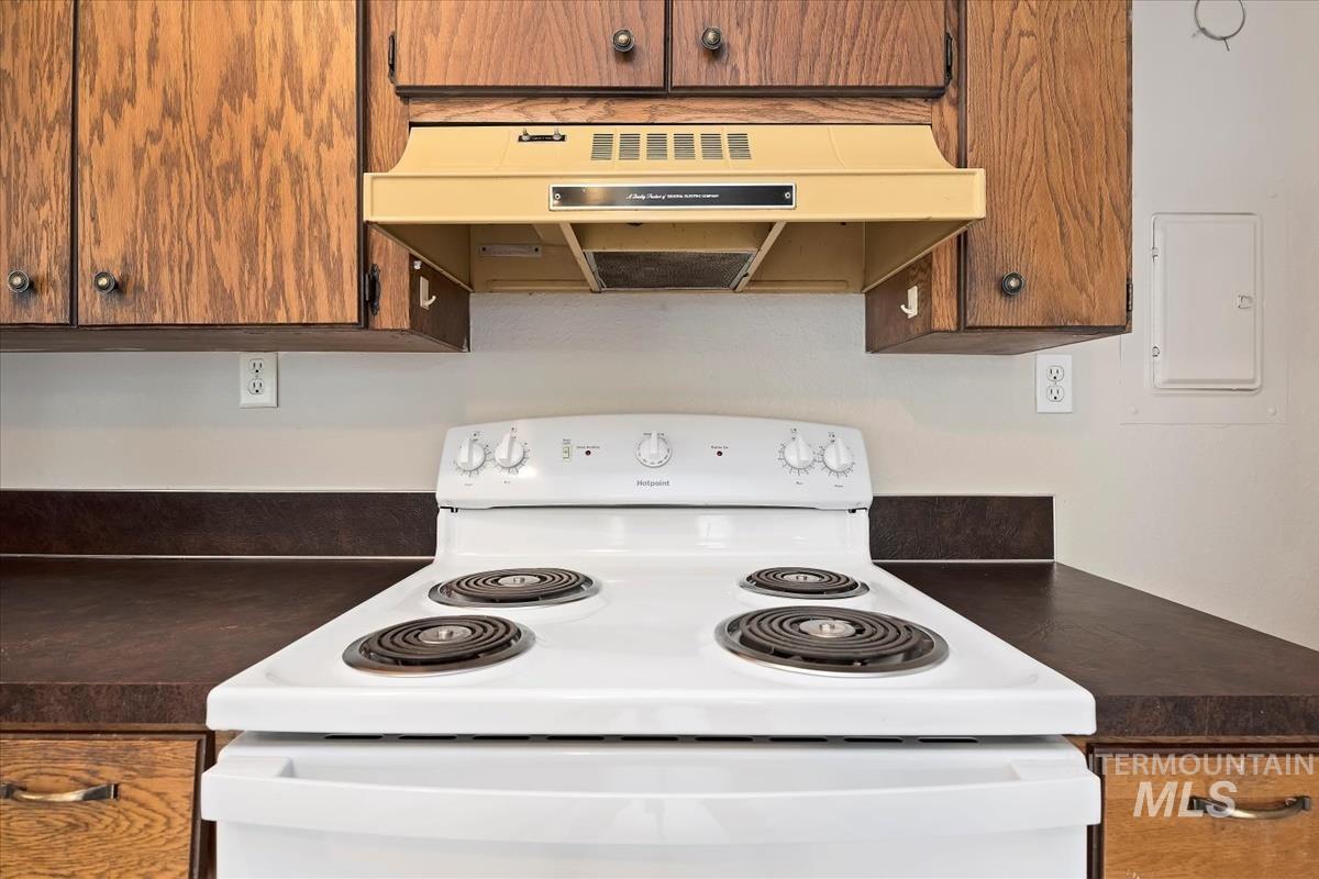 Kitchen featuring dark countertops, white electric stove, under cabinet range hood, brown cabinets, and electric panel