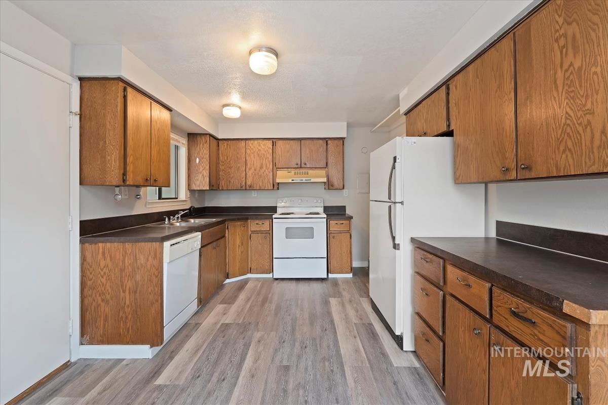 Kitchen with white appliances, light wood-style flooring, dark countertops, and brown cabinetry