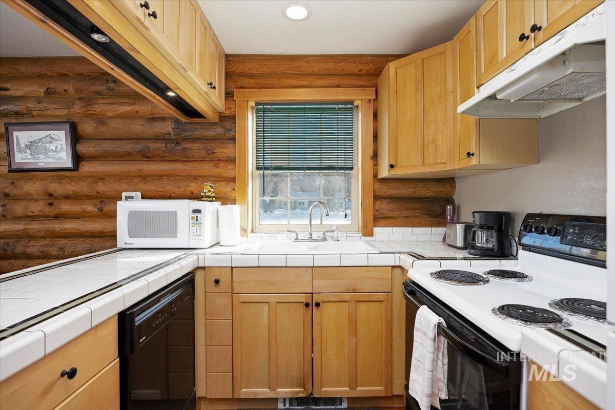 Kitchen with electric range, under cabinet range hood, light brown cabinetry, tile countertops, and log walls