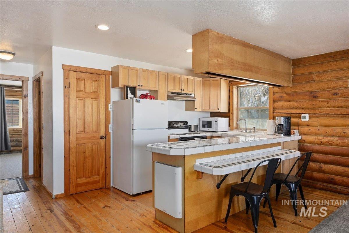 Kitchen with a peninsula, white appliances, light wood-type flooring, a kitchen bar, and tile countertops