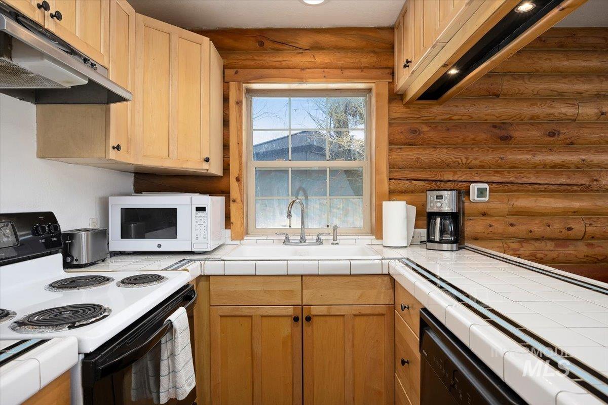 Kitchen with white appliances, under cabinet range hood, log walls, light brown cabinetry, and tile countertops