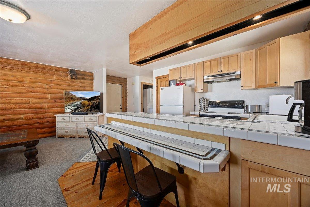 Kitchen featuring light brown cabinets, a kitchen breakfast bar, white appliances, tile countertops, and rustic walls