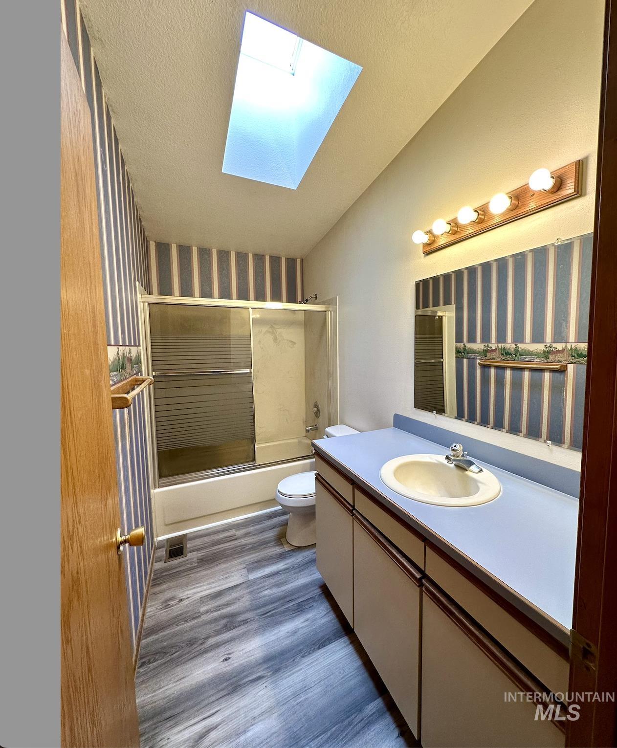 Full bath featuring wallpapered walls, bath / shower combo with glass door, a skylight, vanity, and dark wood-type flooring
