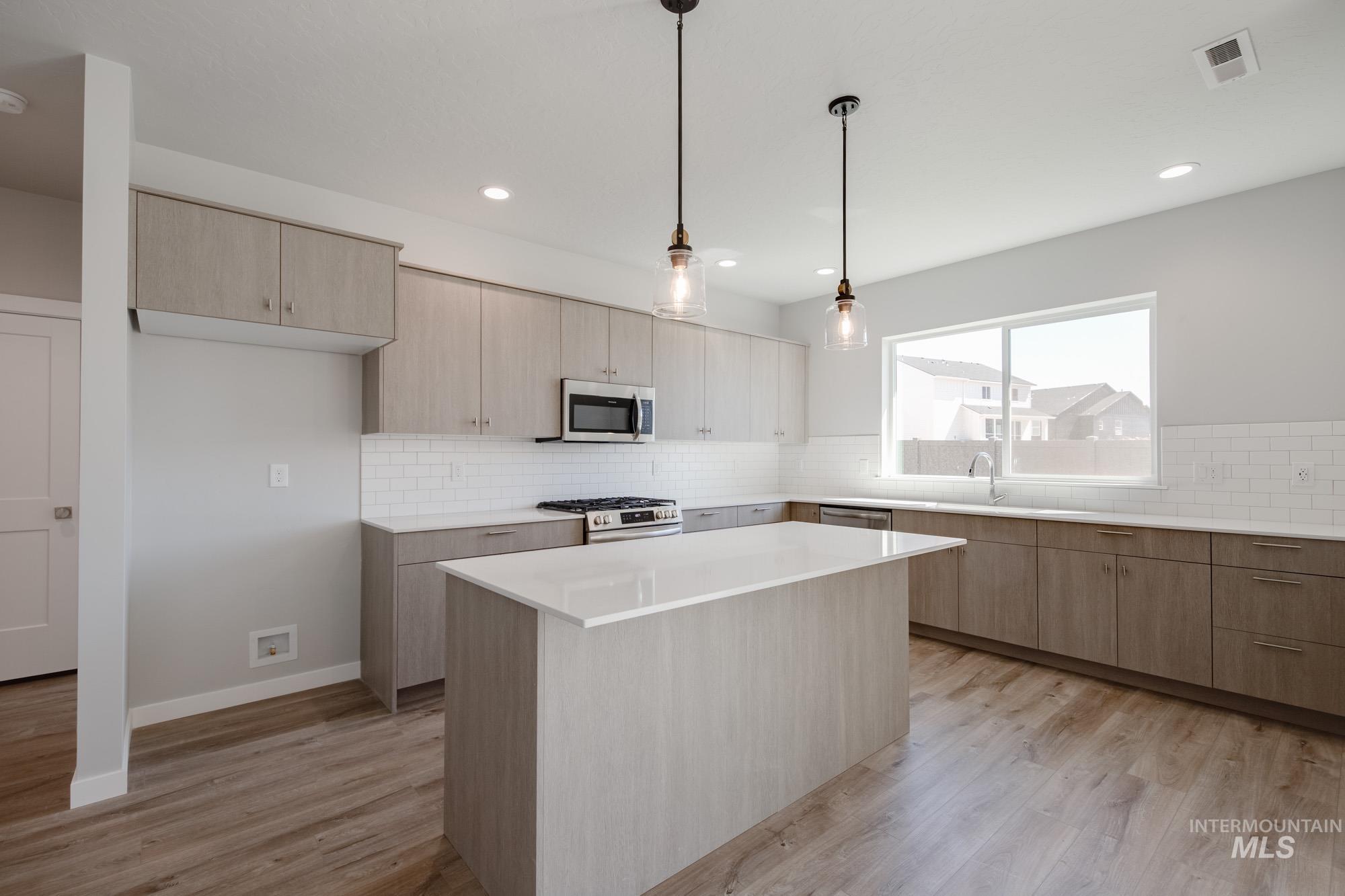Kitchen with light brown cabinetry, hanging light fixtures, a center island, appliances with stainless steel finishes, and light wood-style floors