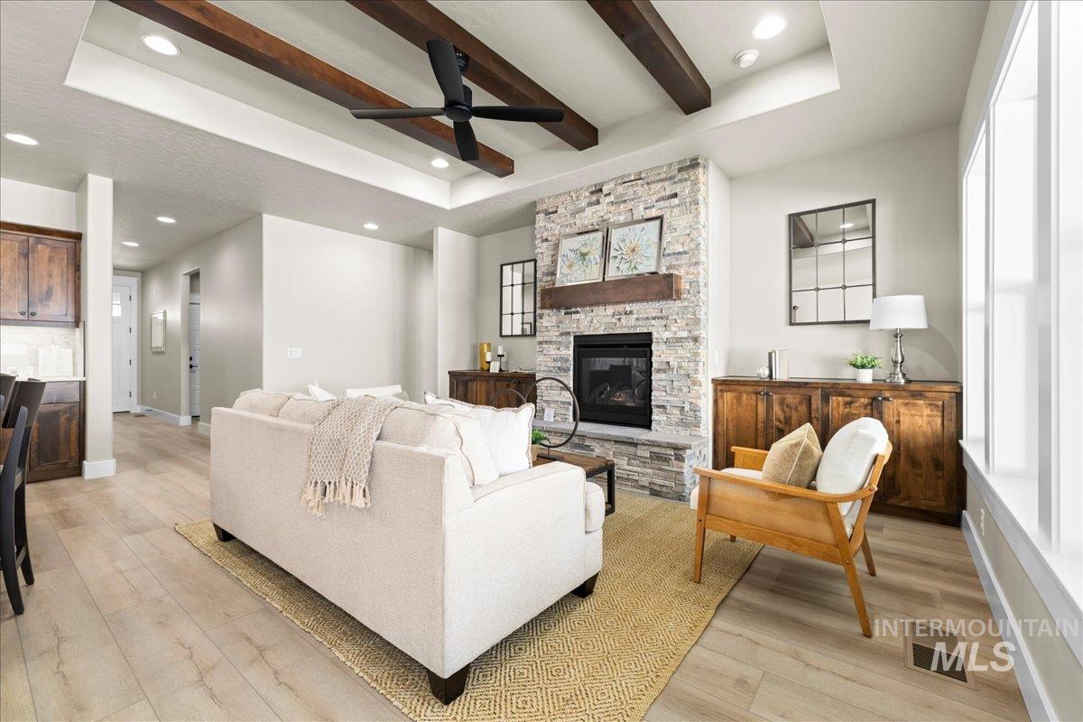 Living area featuring beam ceiling, light wood-style flooring, a tray ceiling, a ceiling fan, and a stone fireplace