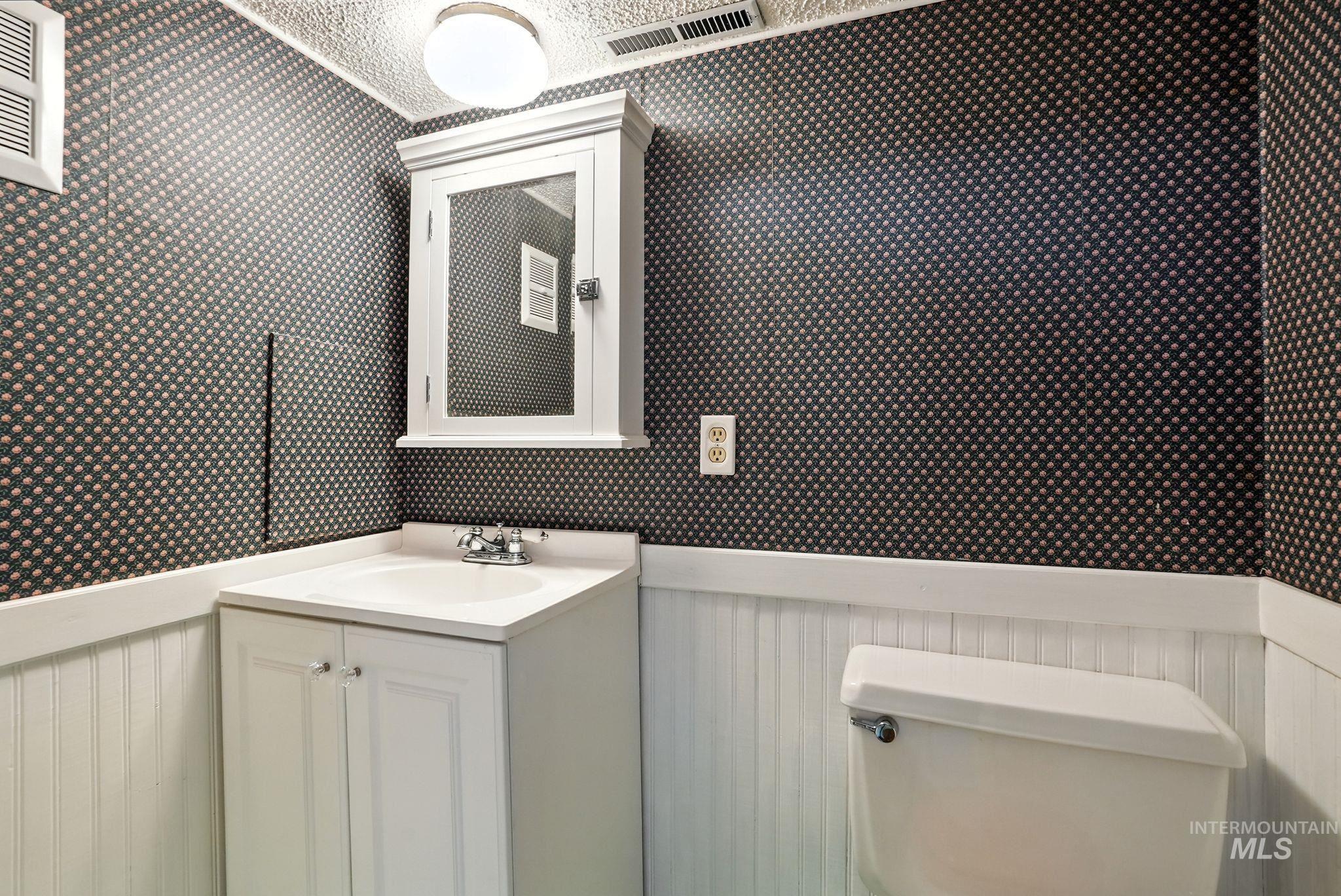 Half bathroom featuring vanity, wallpapered walls, a textured ceiling, and a wainscoted wall
